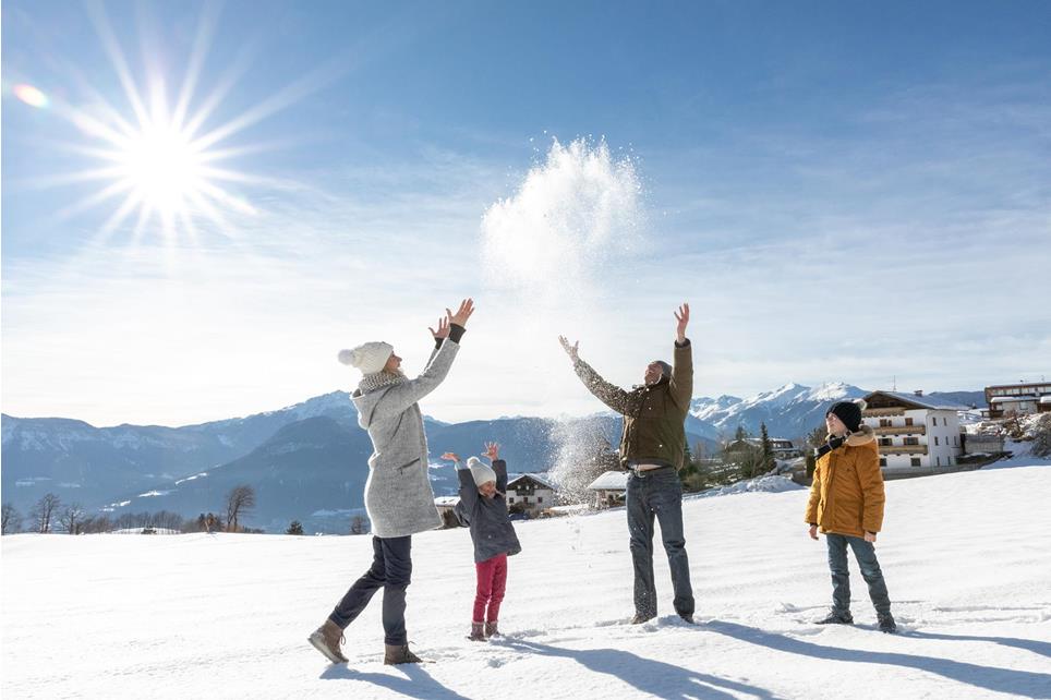 Familie mit Mutter und Vater und zwei Kindern stehen im Schnee im Dorfzentrum von Vöran in Südtirol. Sie werfen den frischen Pulverschnee in die Höhe. Es ist strahlend blauer Himmel und die Sonne scheint.