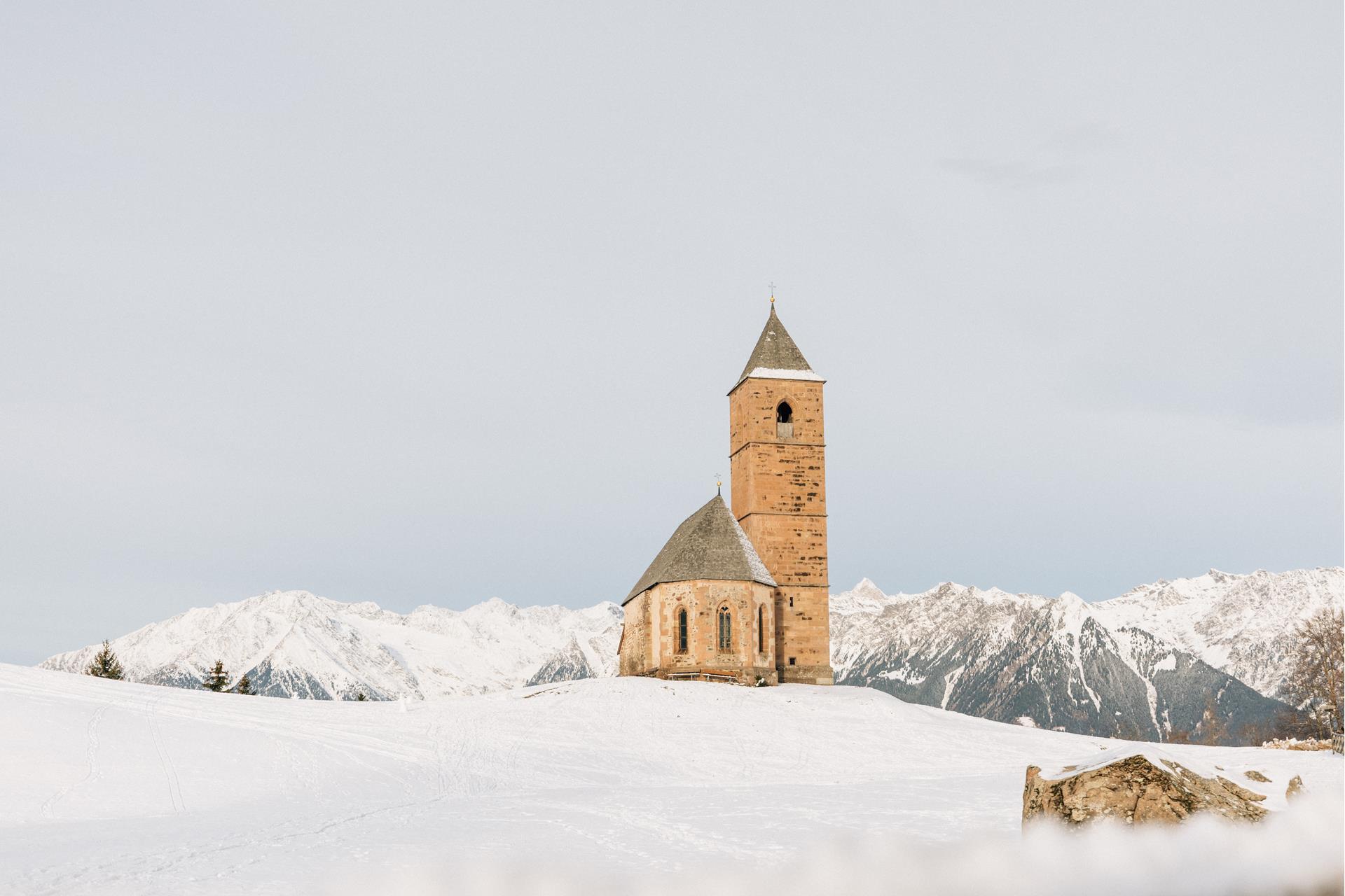 La chiesa di S. Caterina di Avelengo circondata da neve fresca. Sullo sfondo le bianche cime delle montagne in inverno. La suggestiva pietra di fronte alla chiesa.
