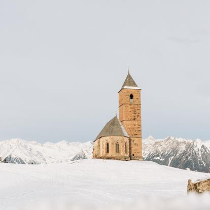 Die St. Kathrein Kirche in der Scharte in Hafling umgeben von frischem pulverschnee. Im Hintergrund die weißen Bergspitzen im Winter. Bisschen vor der Kirche der markante Stein.
