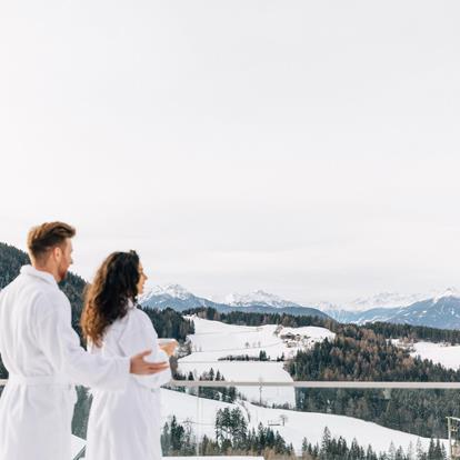 Mann und Frau stehen im Bademantel vor einem Glasbalkon. Aussicht auf die verschneite Winterlandschaft in Hafling, Vöran, Meran 2000