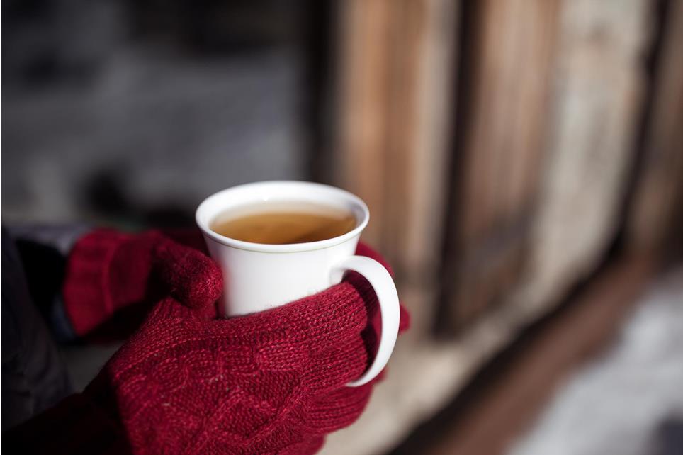 In the center of the picture, there is a white cup of tea held by a person wearing red knit gloves. In the background, you can see the wooden wall of an alpine hut and snow. The image is from Hafling-Verano-Merano 2000.