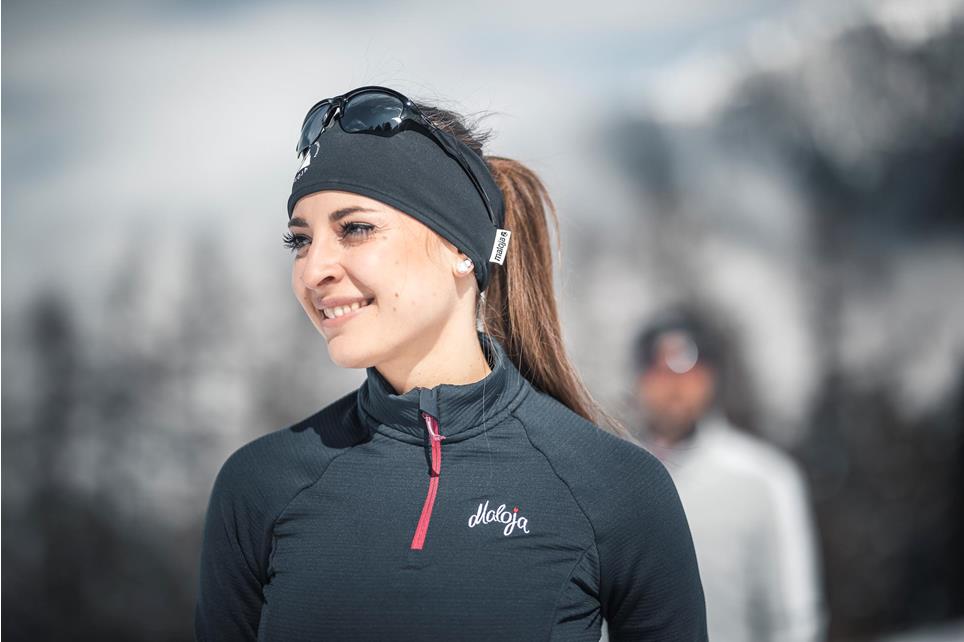 Winter woman in sporty gray attire. She is wearing sunglasses and a functional headband, and her brown hair is in a braid. In the background, the white winter landscape of Hafling-Verano-Merano 2000 in South Tyrol, Italy.