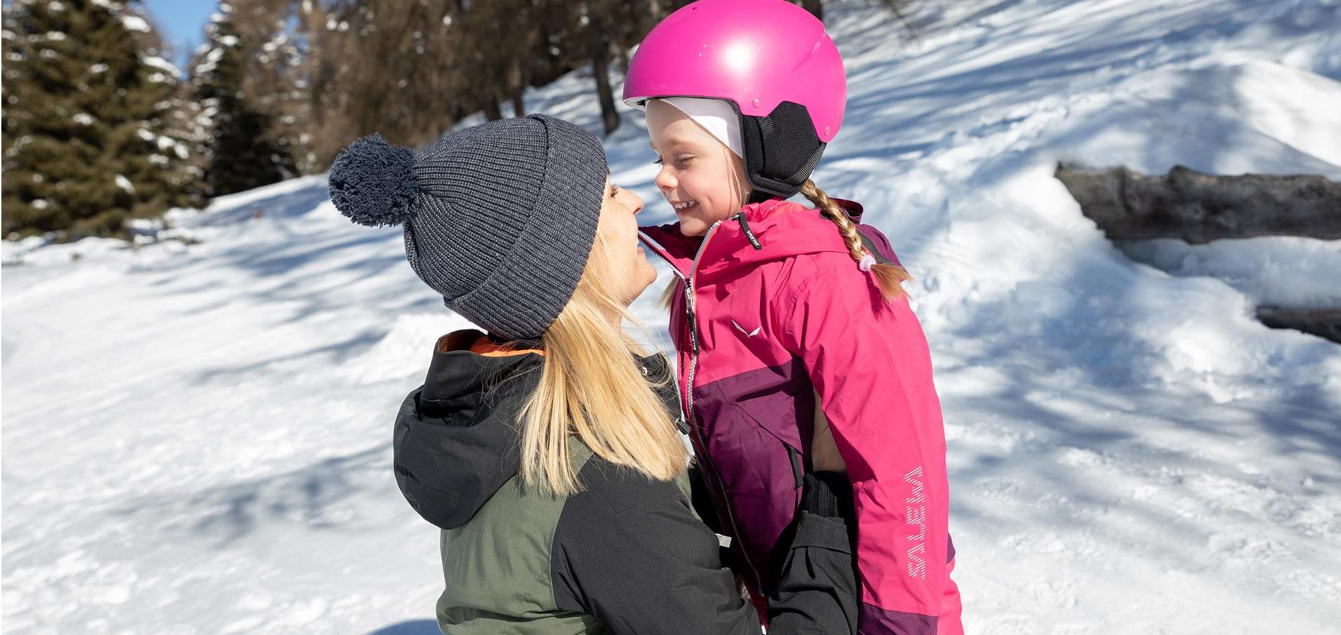 Madre e figlia in inverno a Verano, in Trentino-Alto Adige. Entrambe ridono e si guardano. La bambina ha circa 4 anni e indossa una tuta da sci rosa con un casco rosa.