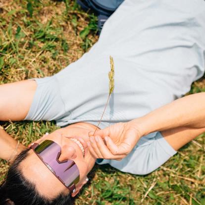 Woman with blue T-shirt and violet biz glasses lies on green meadow and has blade of grass in her mouth.