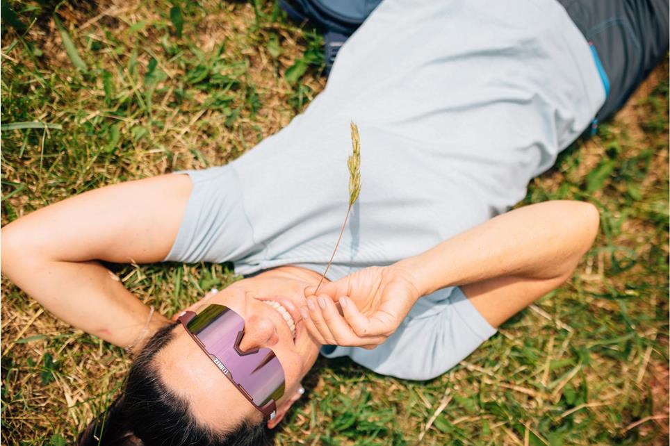 Woman with blue T-shirt and violet biz glasses lies on green meadow and has blade of grass in her mouth.