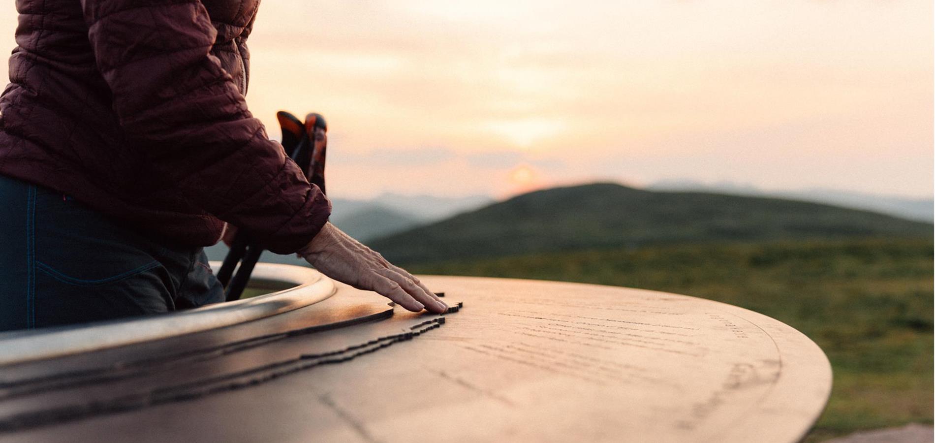 Sunrise at the Kreuzjoch viewing platform. A woman gives her hand to the viewing plate, which is made of rusty iron and shows the mountains surrounding Merano and the Dolomites.