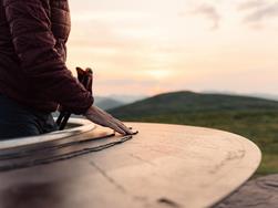 Sunrise at the Kreuzjoch viewing platform. A woman gives her hand to the viewing plate, which is made of rusty iron and shows the mountains surrounding Merano and the Dolomites.